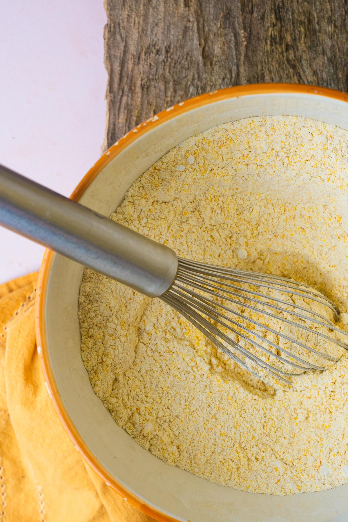 A metal whisk resting in a bowl of yellow cornmeal on a wooden surface. An orange cloth is partially visible nearby.