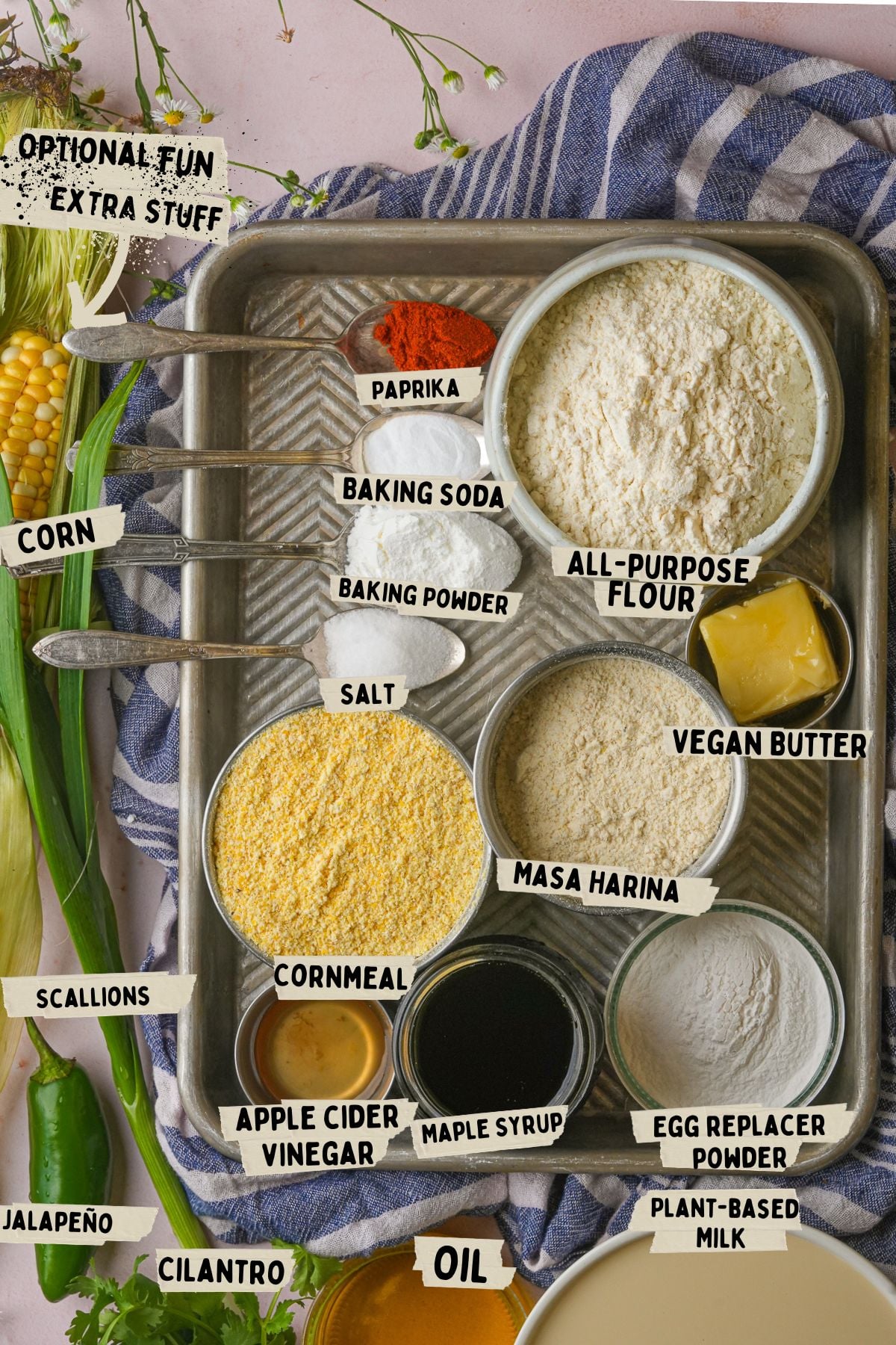 Ingredients for a vegan cornbread recipe including cornmeal, all-purpose flour, masa harina, vegan butter, maple syrup, and various seasonings, arranged on a baking tray with optional add-ons like jalapeño and cilantro.