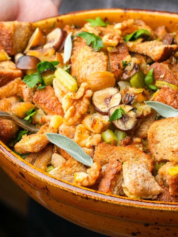 A close-up of a bowl of vegan stuffing with bread cubes, celery, herbs, nuts, and slices of mushrooms, being held by two hands.