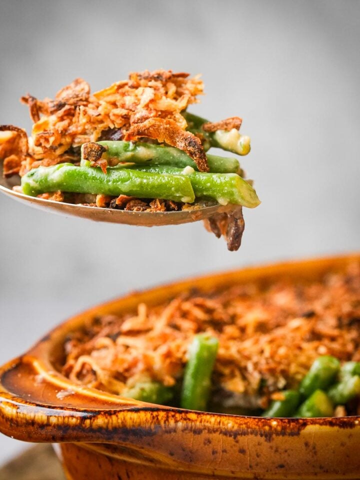 A spoon lifting a serving of green bean casserole topped with crispy fried onions from a brown baking dish.