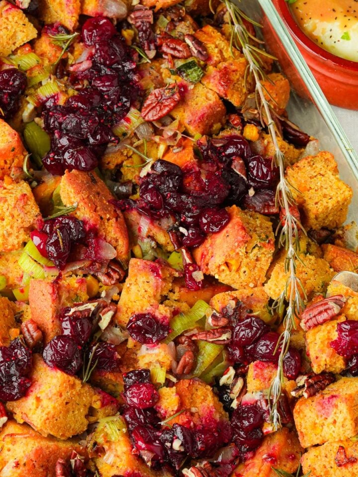 A baking dish filled with vegan cornbread stuffing, featuring chunks of cornbread, cranberries, pecans, and celery. A metal serving spoon rests on the dish. A small bowl with dipping sauce is nearby.