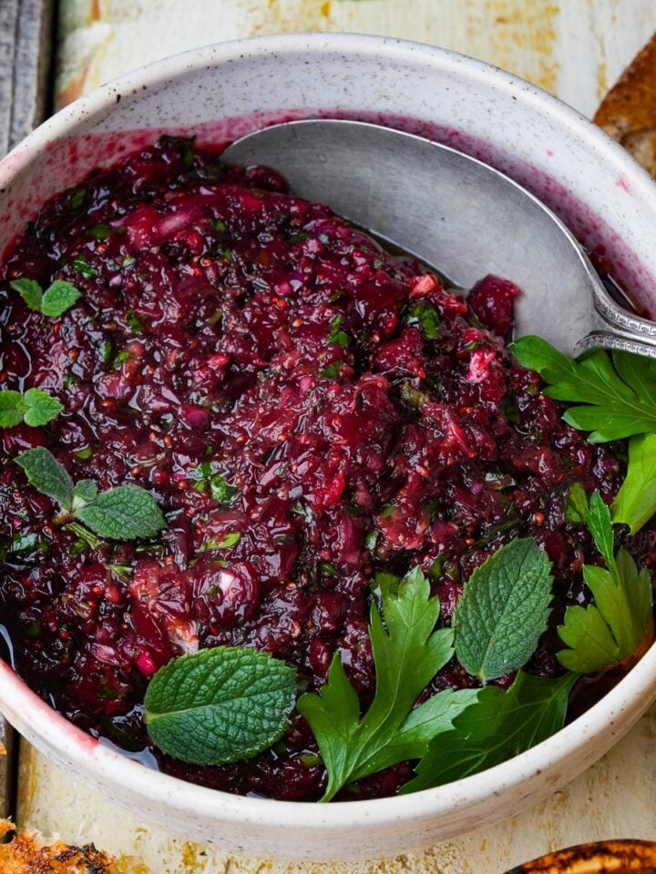 A bowl of red Cranberry Ezme garnished with fresh mint and parsley, with a spoon on the side. Toasted bread pieces are visible nearby.
