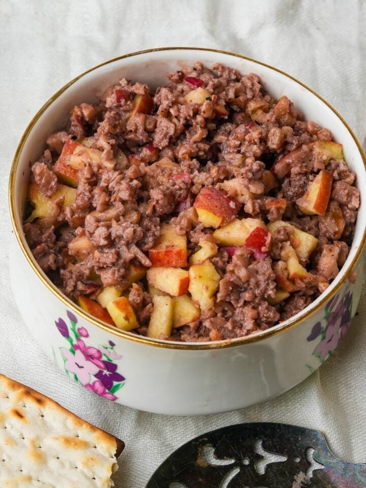 A bowl filled with charoset made from apples and minced nuts, resting on a white cloth. A cracker and a silver utensil are nearby.