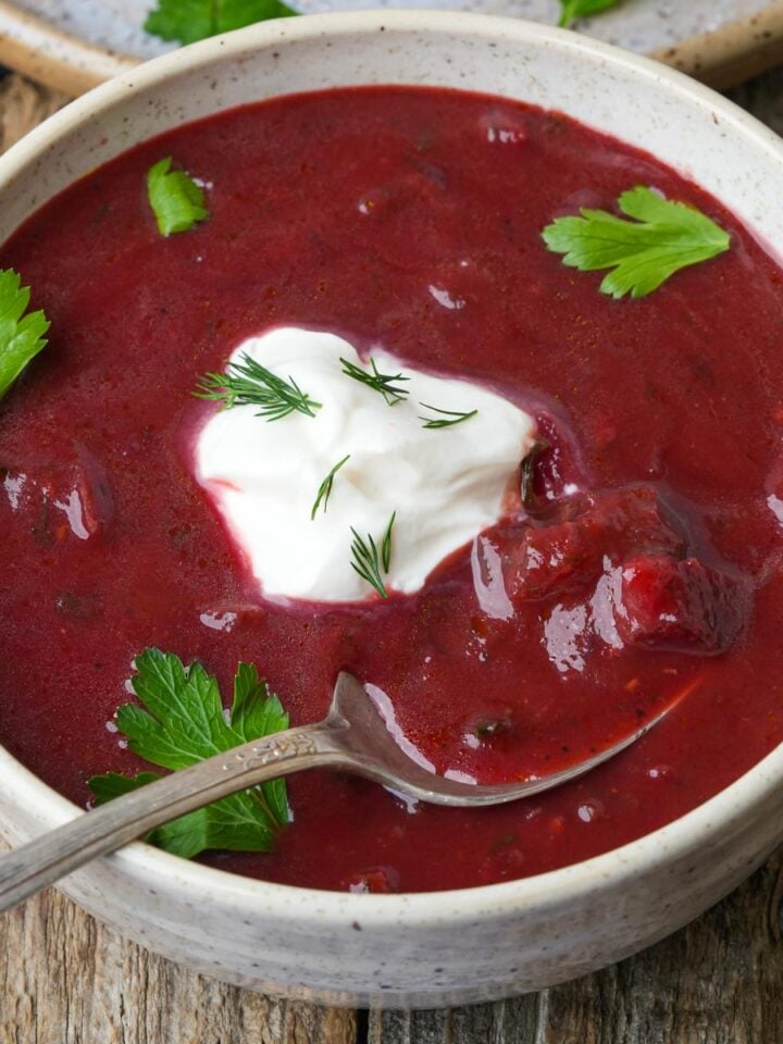 A bowl of red vegan borscht soup with a dollop of tofu sour cream and dill on top. Garnished with parsley leaves, and a spoon rests inside the bowl.