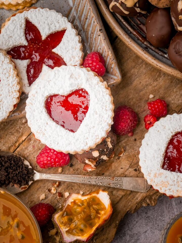 Cookies filled with red jam in heart and star shapes, surrounded by raspberries, chocolate truffles, a spoon with cocoa powder, and a halved passion fruit on a wooden surface.