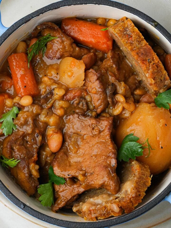 Vegan cholent in a round blue pot containing seitan, carrots, potatoes, beans, and garnished with parsley.