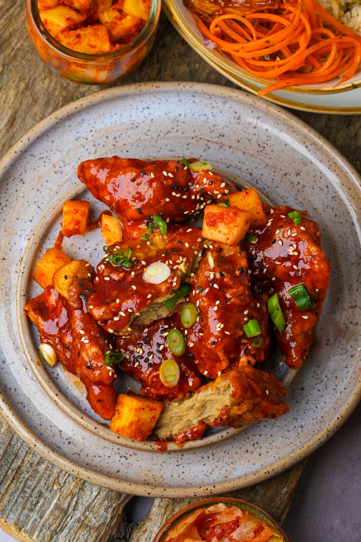 A plate of spicy glazed vegan Korean fried chicken garnished with green onions and sesame seeds, accompanied by cubed potatoes.