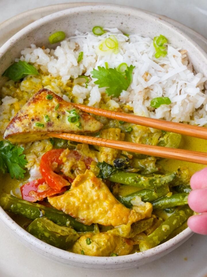 A bowl of green curry tofu served with white rice. A hand holding chopsticks is picking up a piece of tofu.