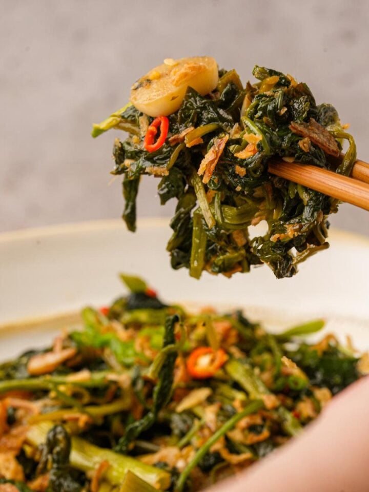 Chopsticks holding a portion of stir-fried rau muong with garlic and chili, above a plate of the same dish.