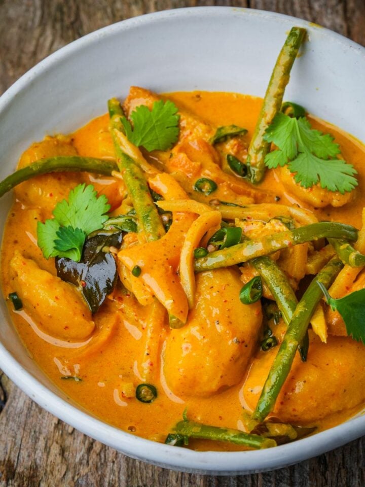 A bowl of vegan yellow curry with chunks of tofu, green beans, sliced onions, and cilantro leaves on a wooden surface.
