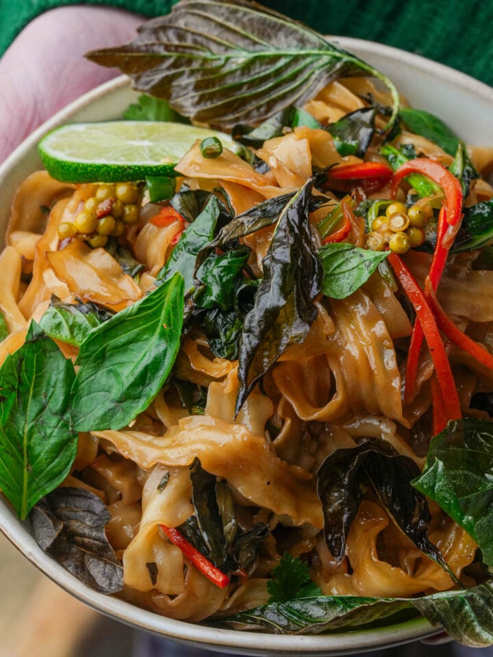 A bowl of stir-fried noodles with fresh basil leaves, red bell pepper strips, a lime wedge, and garnished with green peppercorns and herbs.