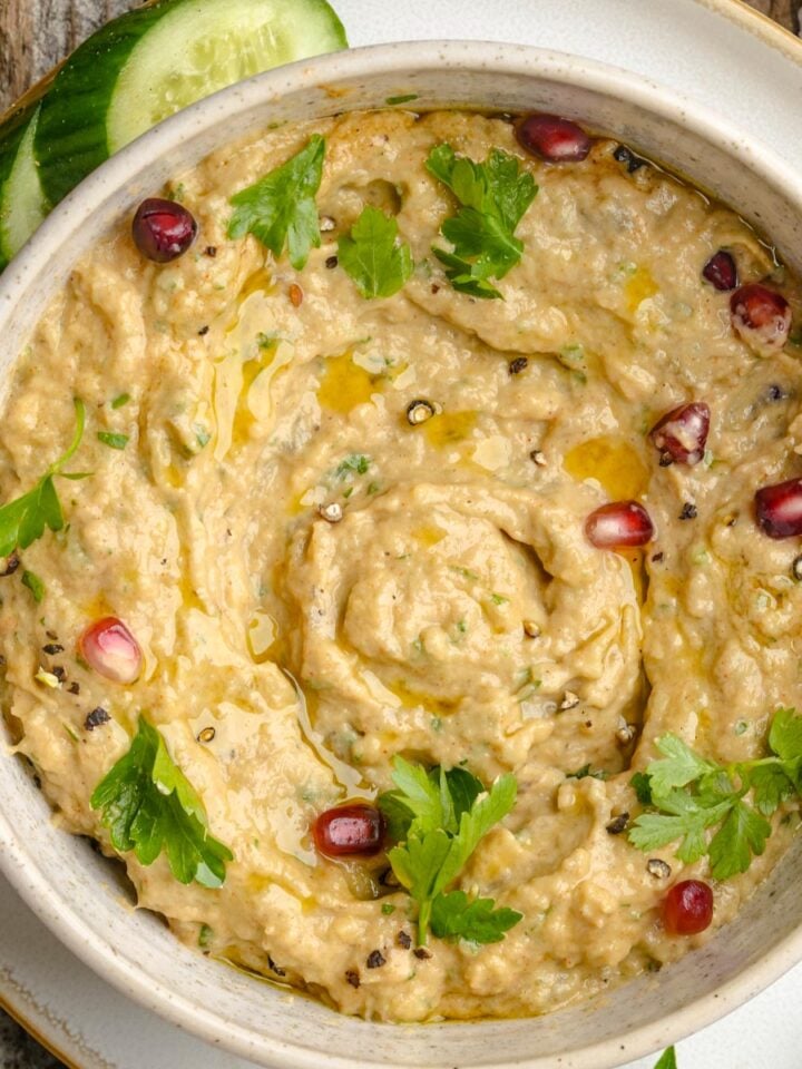 A bowl of Lebanese baba ganoush garnished with parsley, pomegranate seeds, black pepper, and olive oil, with cucumber slices on the side.