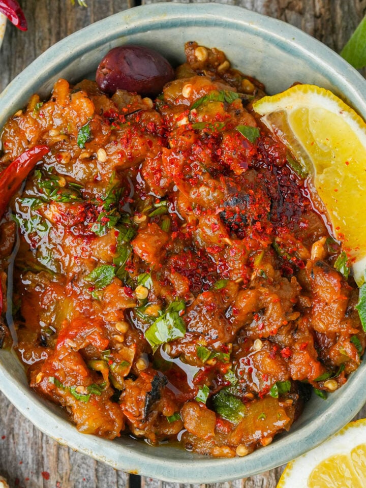 A bowl of zaalouk garnished with fresh herbs, a dried red chili, olives, and a lemon wedge, placed on a rustic wooden surface.