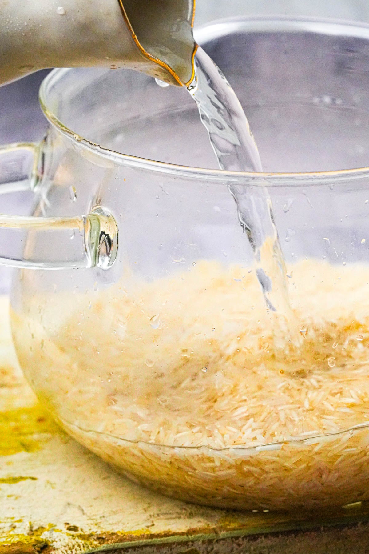 Clear glass pot filled with uncooked rice as water is being poured in from a jug, set on a countertop.
