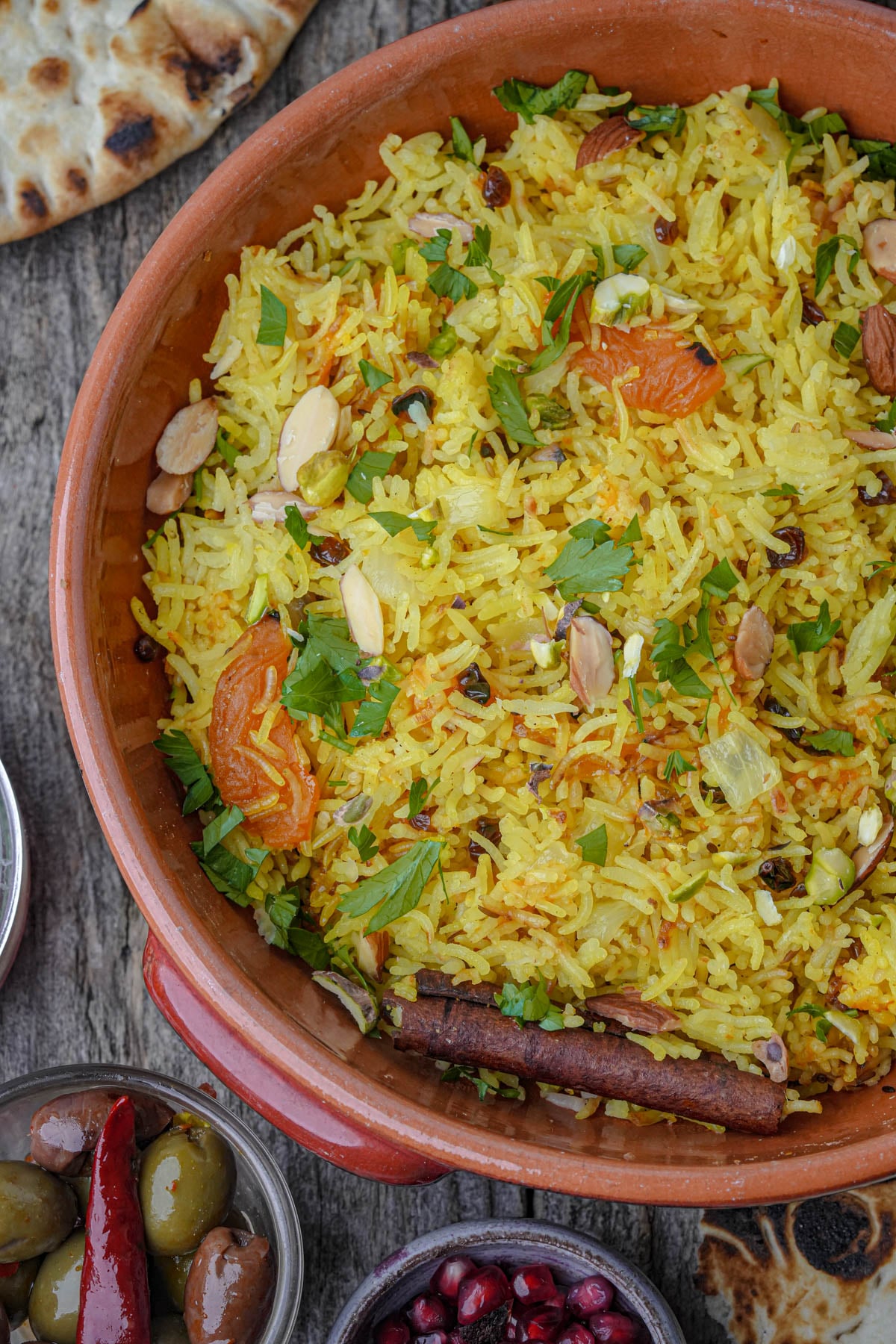 A bowl of Moroccan rice garnished with nuts, dried fruits, and herbs, surrounded by sides including flatbread, olives, and pomegranate seeds.