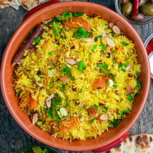 A ceramic dish filled with Moroccan rice garnished with sliced almonds, fresh herbs, and dried fruit, with pieces of naan bread and small bowls of condiments nearby.