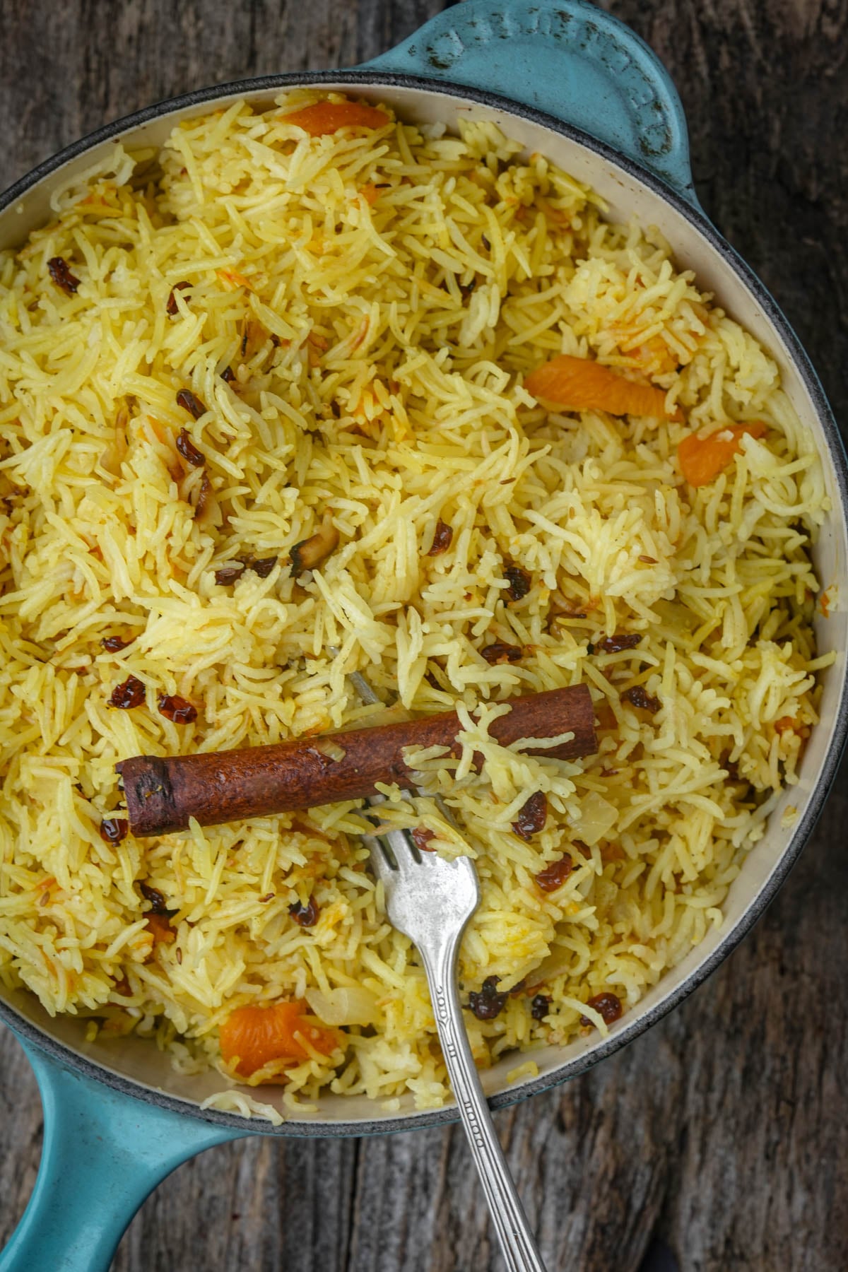 A pot of Moroccan rice with raisins, apricot pieces, and a cinnamon stick, with a fork resting inside; pot sits on a rustic wooden surface.