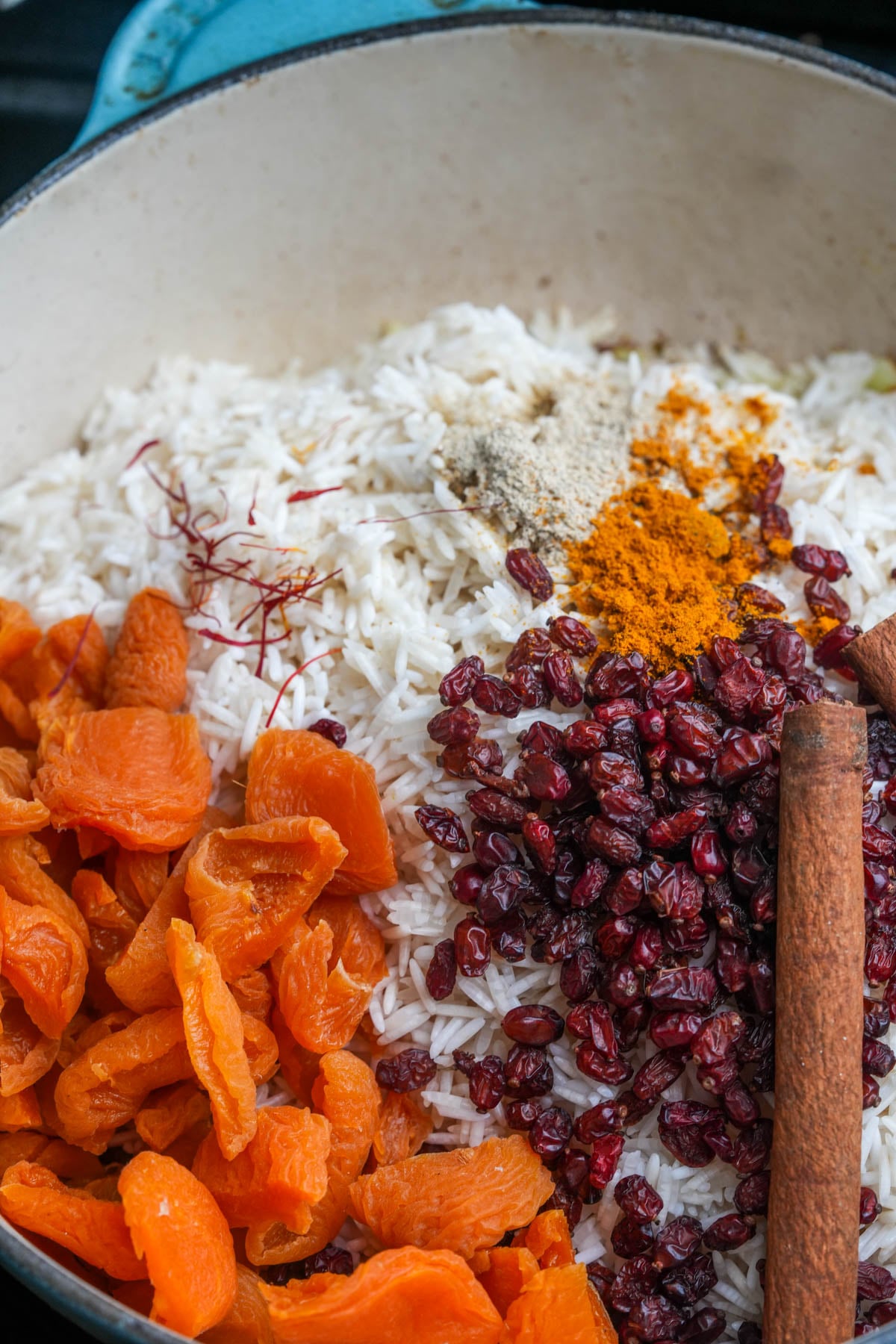A pot filled with uncooked Moroccan rice, dried apricots, dried barberries, whole cinnamon sticks, and assorted ground spices, including turmeric and saffron threads.
