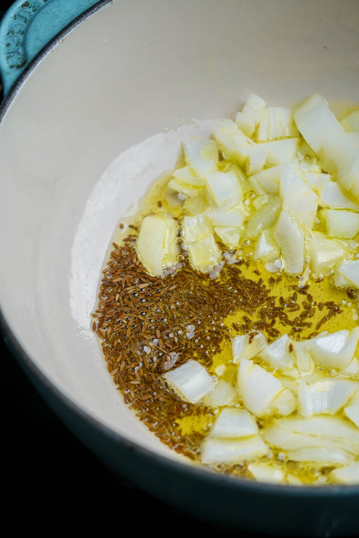 Chopped onions and cumin seeds are sautéing in oil in a light-colored pot.