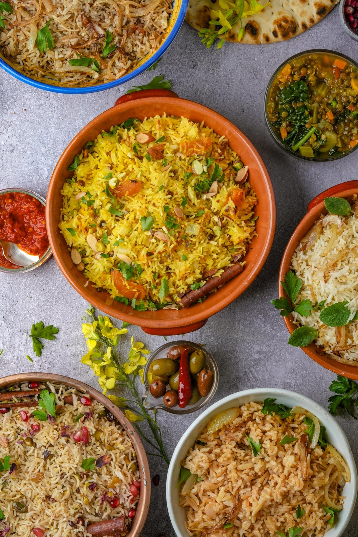 A variety of rice dishes in bowls, including Persian rice, Moroccan rice, Egyptian rice, Lebanese rice, and Turkish rice, garnished with herbs and pomegranate seeds, surrounded by small bowls of olives, harissa, and other side dishes.