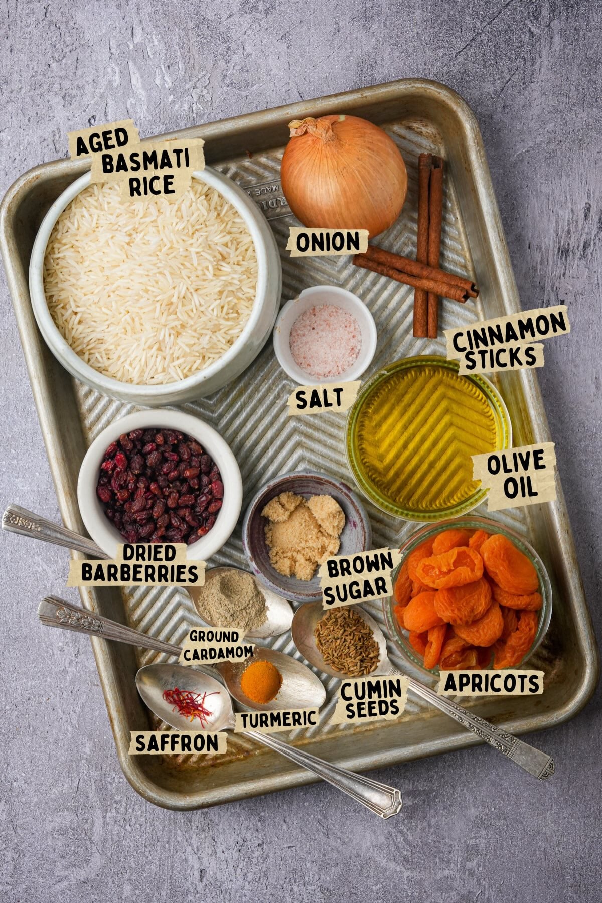 A baking tray with bowls and spoons holding ingredients for a Moroccan rice recipe: aged basmati rice, onion, cinnamon sticks, olive oil, salt, dried barberries, brown sugar, apricots, cumin seeds, turmeric, cardamom, and saffron.