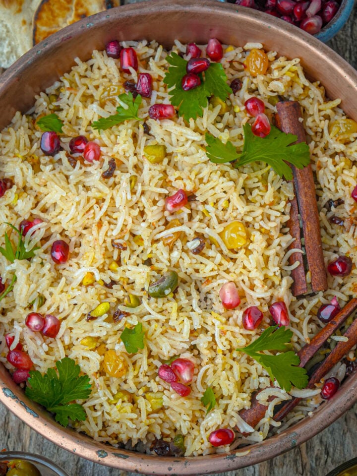 A bowl of Persian rice garnished with pomegranate seeds, cilantro, whole cinnamon sticks, raisins, and pistachios.