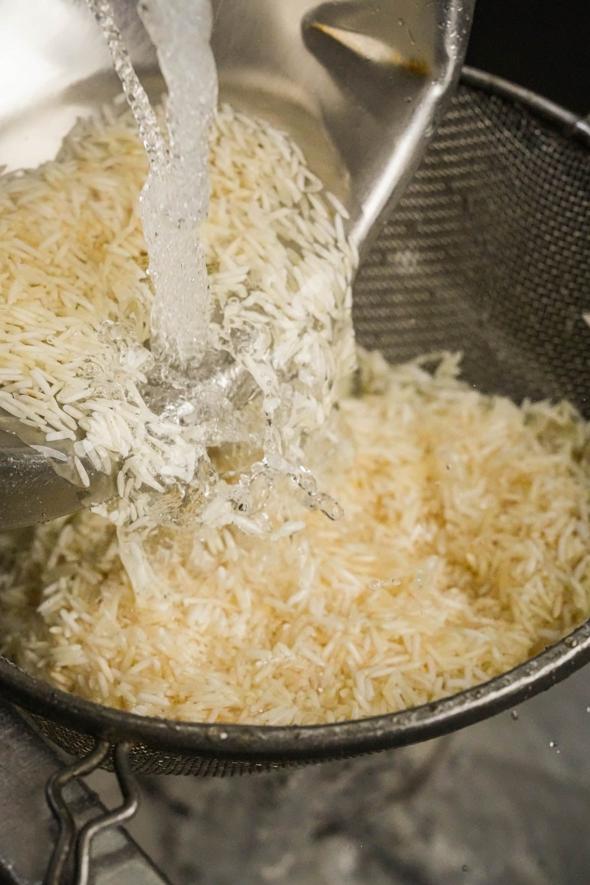 Uncooked rice being rinsed under running water in a metal strainer.