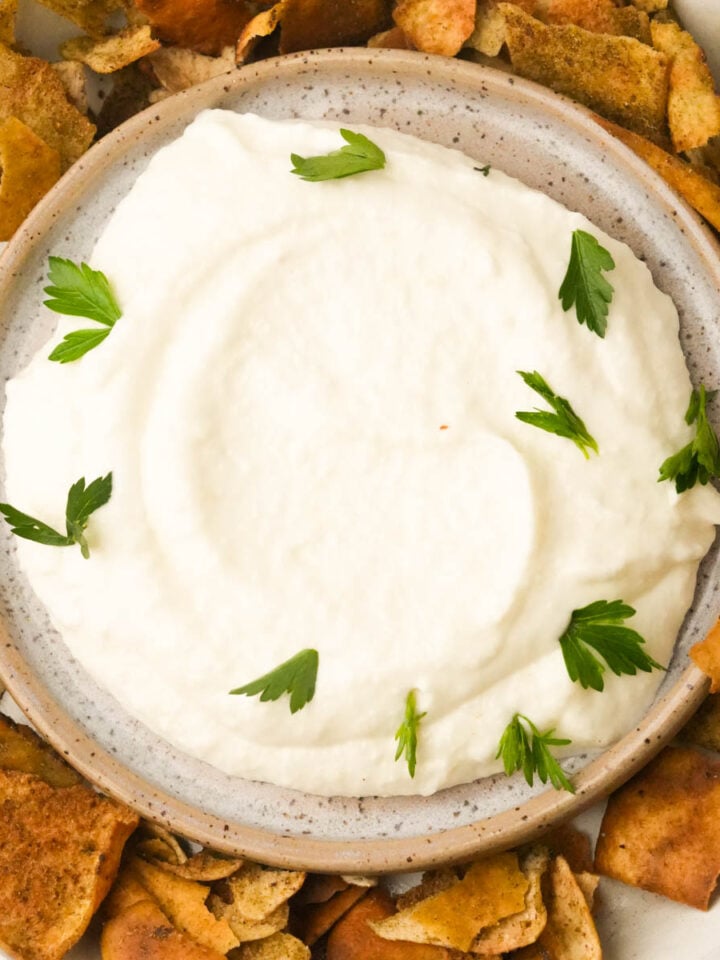 A bowl of creamy white toum garnished with parsley, surrounded by pieces of toasted pita chips.