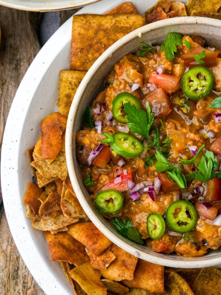 Bowl of chunky ful medames garnished with sliced jalapeños, red onion, parsley, and tomatoes, surrounded by pita chips on a white plate. Lemon and garlic are nearby on a wooden surface.