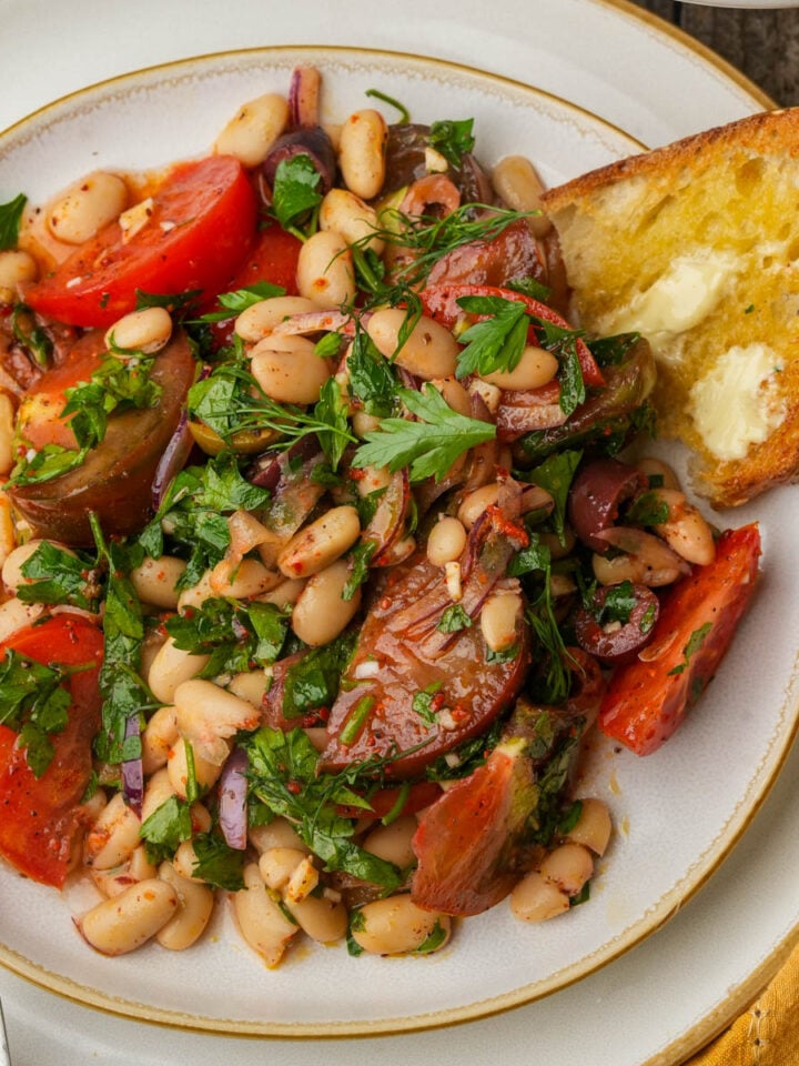A plate of piyaz salad with tomatoes, red onion, parsley, and olives, served with a slice of buttered toasted bread and a fork on the side.