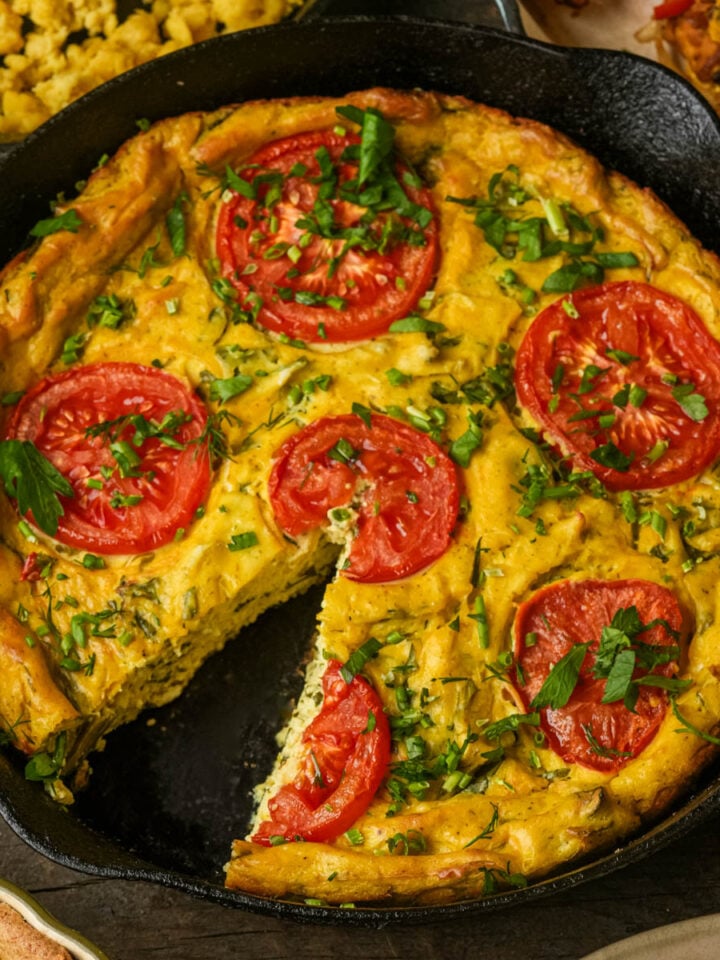 A baked vegan frittata in a cast iron skillet, topped with sliced tomatoes and herbs, with one slice removed.
