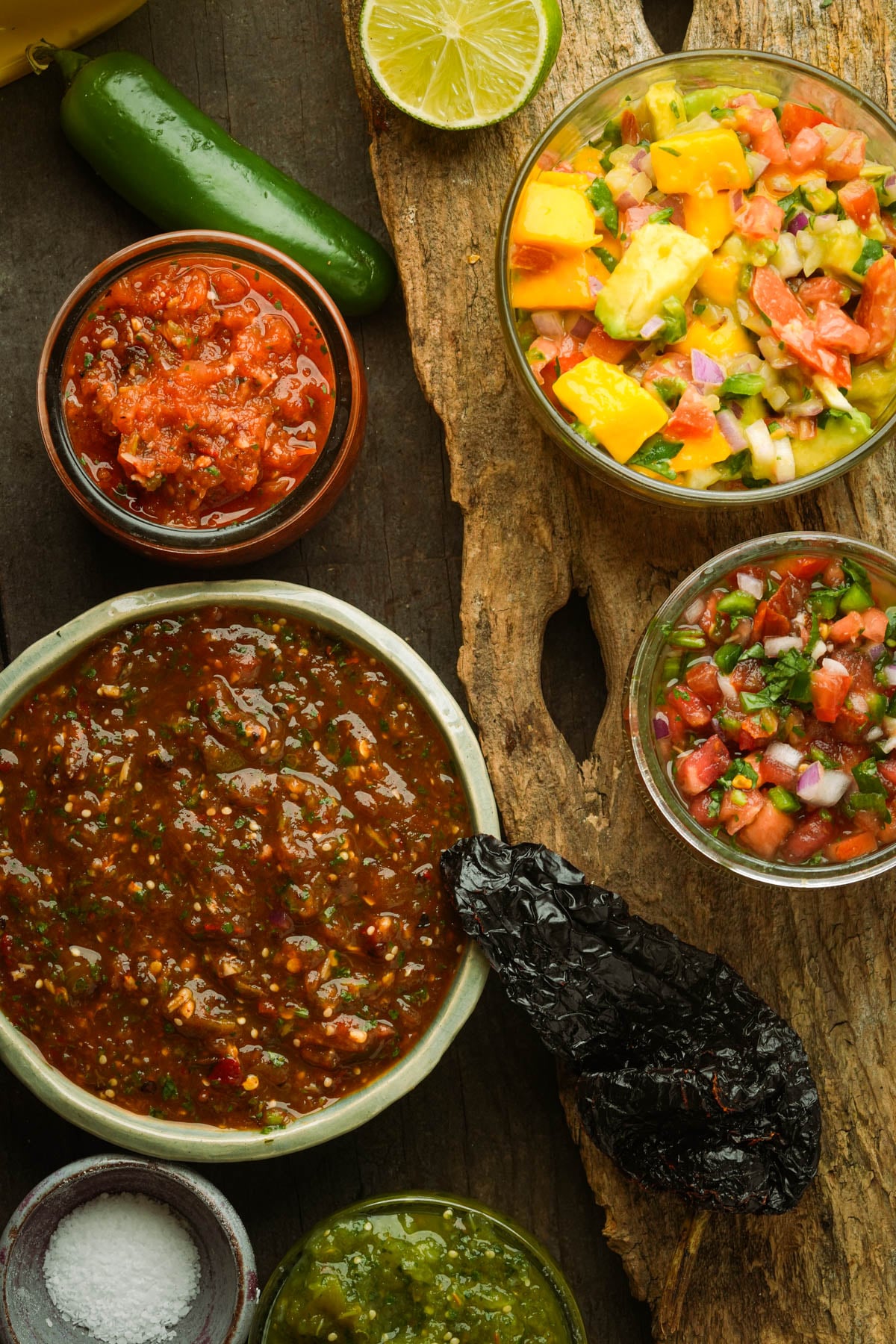 Assorted salsas in small bowls, including salsa fresca, jalapeno salsa, mango salsa, chile de arbol salsa, and salsa ranchera, arranged on a wooden surface with a chili pepper, salt, and dried pepper.