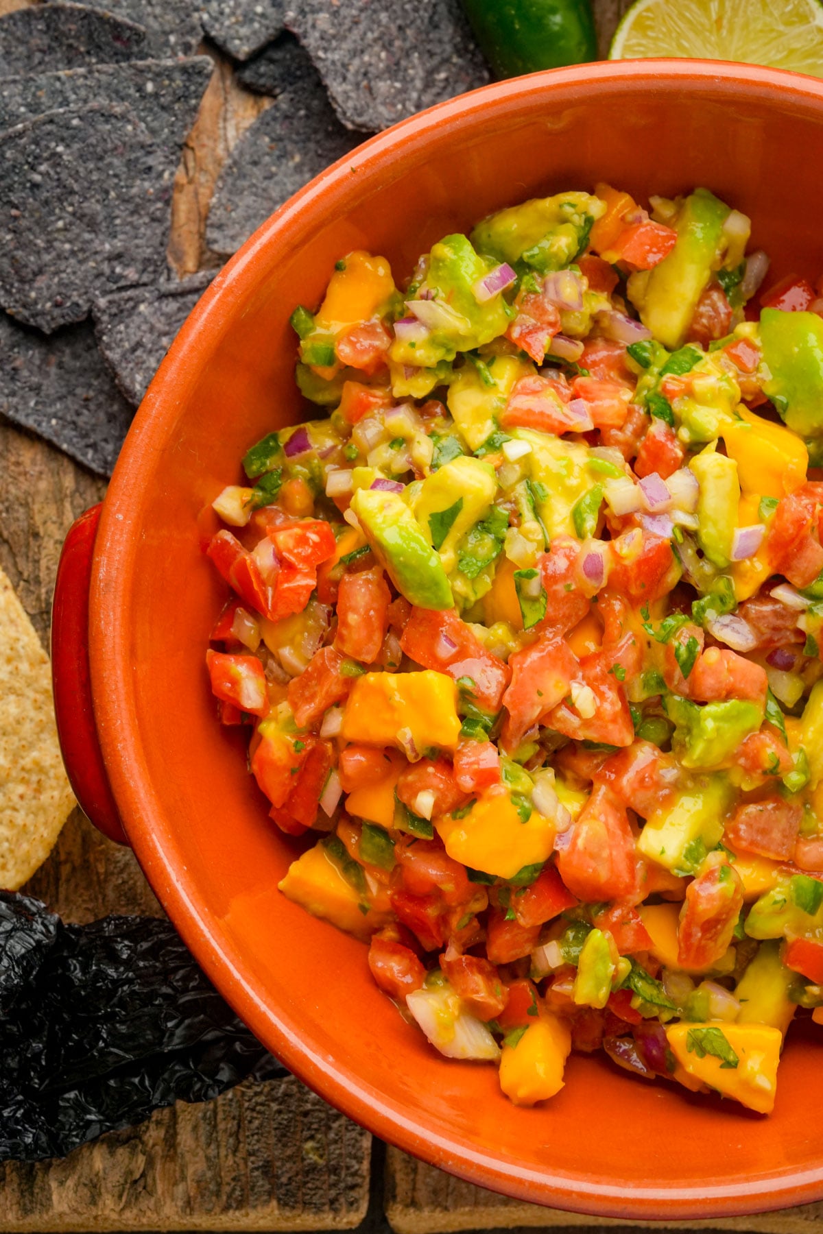 A bowl of chunky mango salsa with diced avocado, tomato, red onion, and herbs, surrounded by blue corn tortilla chips on a wooden surface.