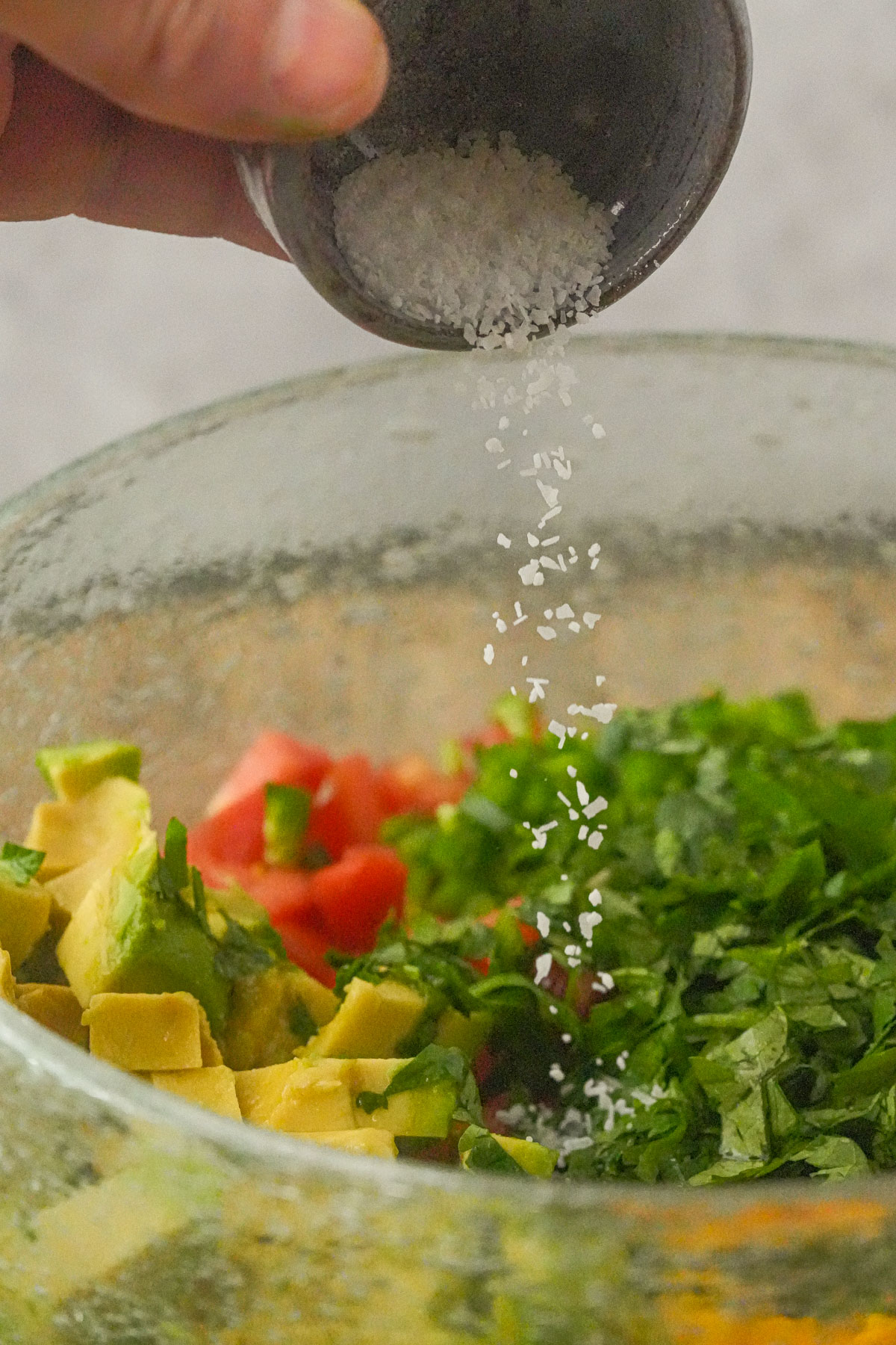 A hand sprinkles salt from a small bowl onto chopped avocado, tomato, and cilantro in a glass mixing bowl.