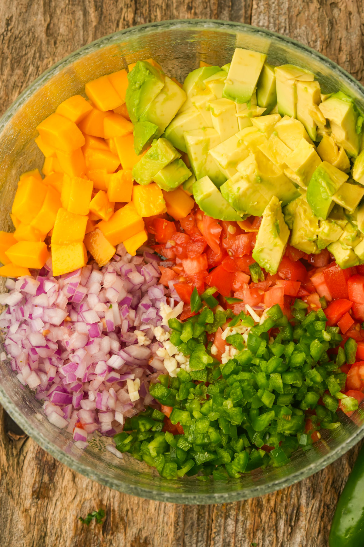 A glass bowl containing diced avocado, mango, tomato, red onion, and jalapeño, arranged in separate sections on a wooden surface.
