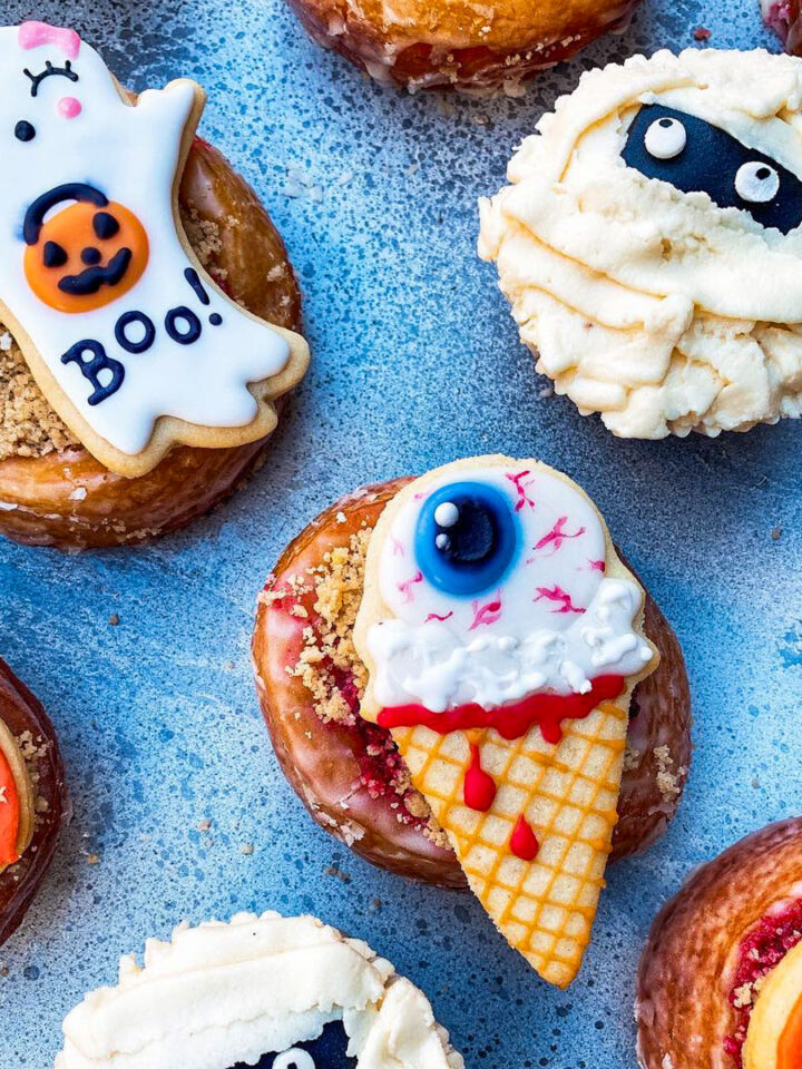 Assorted Halloween-themed donuts decorated with icing shaped like a ghost, mummy, pumpkin, and an eyeball on a cone, arranged on a blue surface.