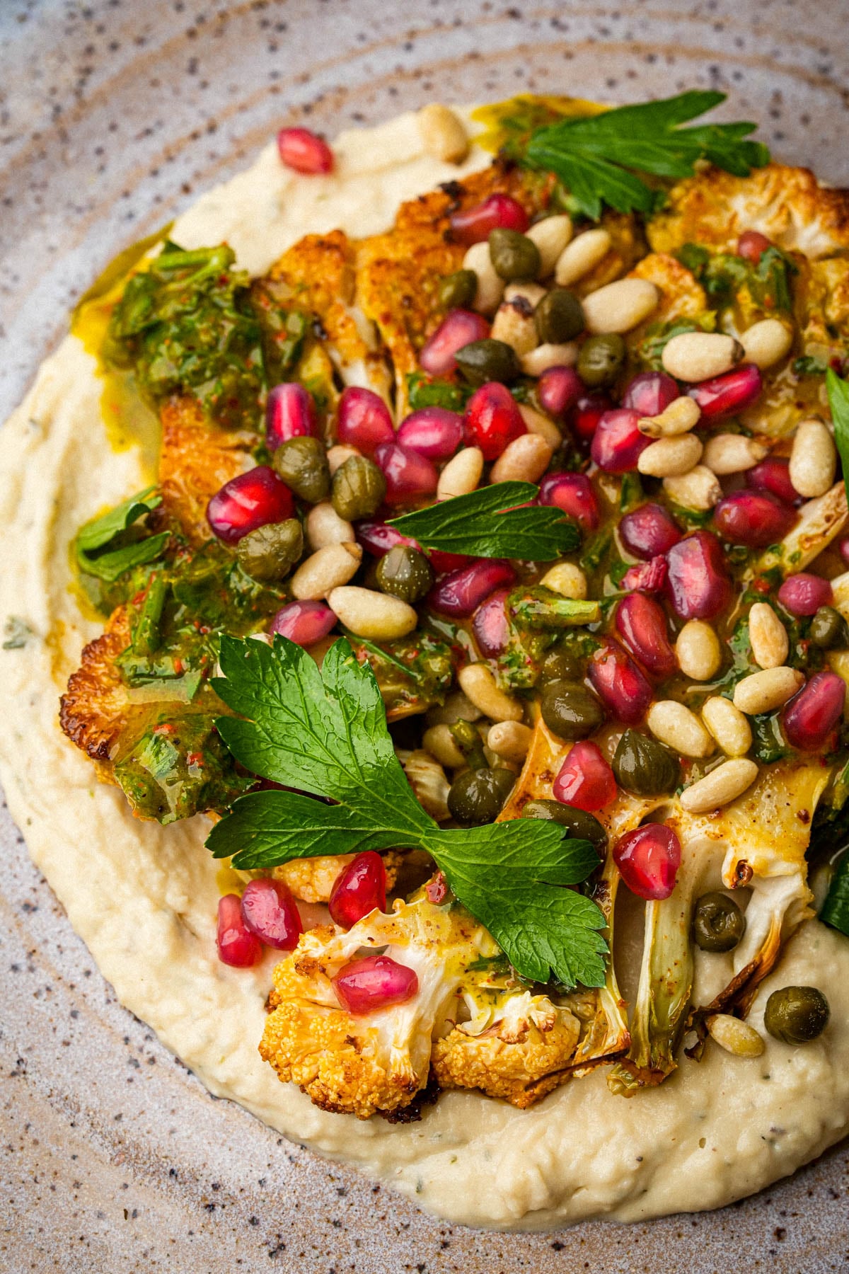 Roasted cauliflower steak on a bed of creamy bean puree, topped with parsley leaves, pomegranate seeds, capers, pine nuts, and a drizzle of green herb sauce, served on a rustic plate.