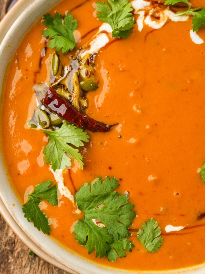 A bowl of creamy orange Thai pumpkin curry soup garnished with fresh cilantro, a swirl of coconut milk, fried chili, seeds, and spices, set on a rustic wooden surface.