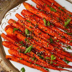 A plate of maple roasted carrots garnished with sesame seeds, chopped chives, and fresh parsley, arranged neatly on a decorative white platter.