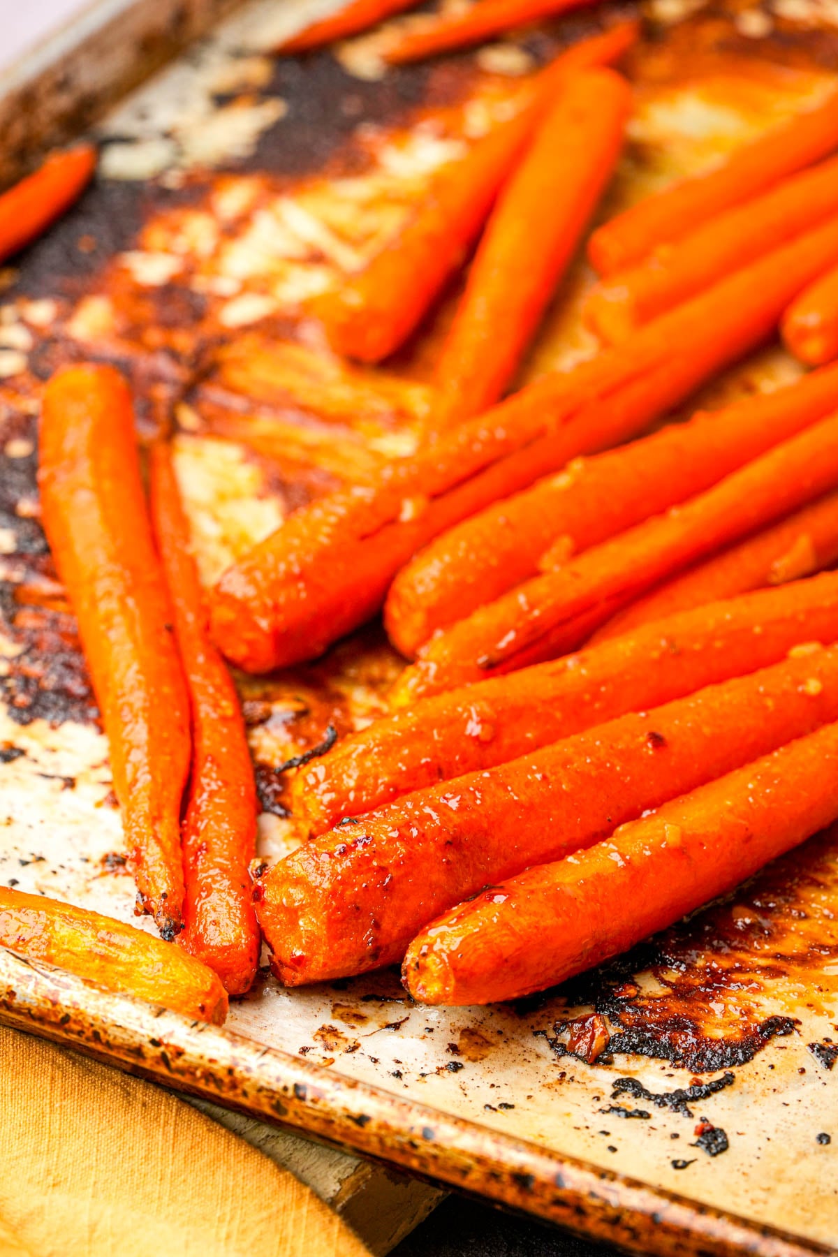 Whole maple roasted carrots coated in a shiny glaze, arranged on a well-used, slightly browned baking sheet. The carrots are caramelized and appear tender, ready to serve.