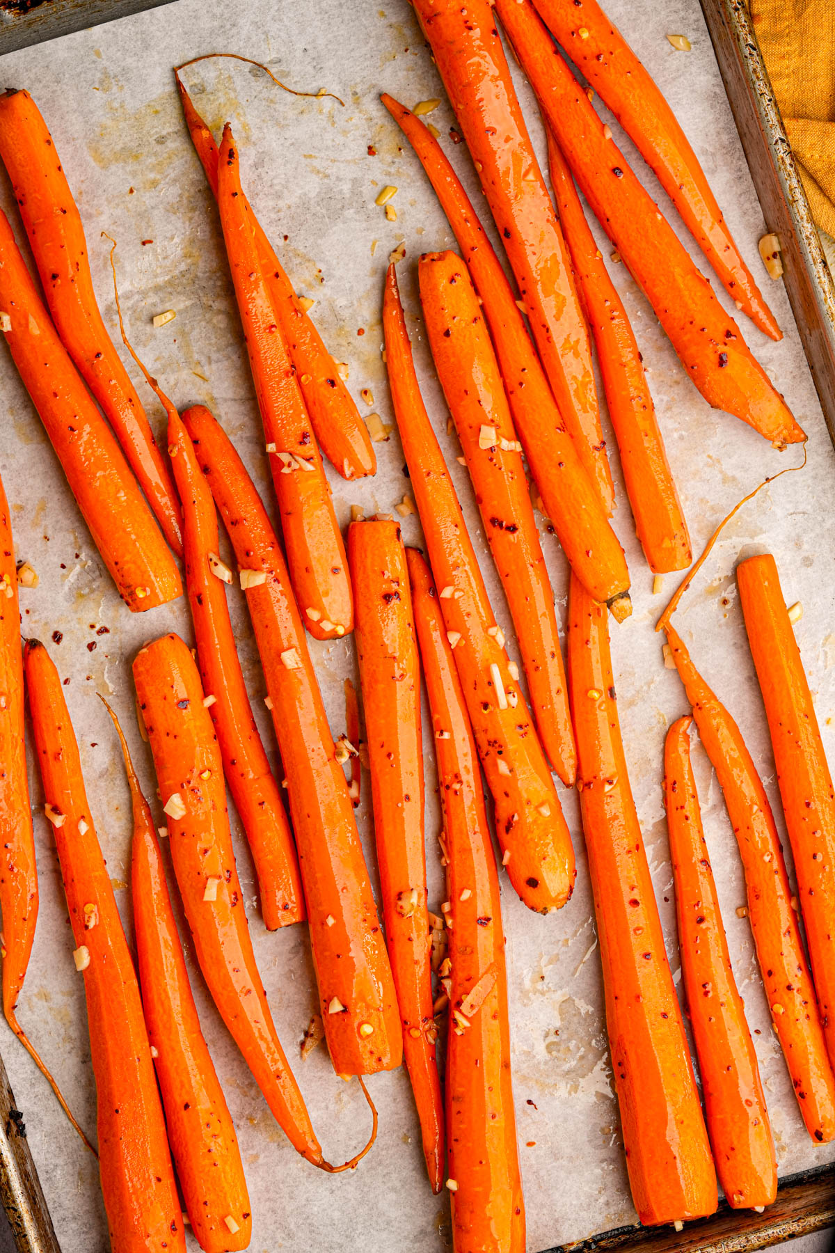 A baking sheet lined with parchment paper holds marinated carrot, seasoned with visible bits of minced garlic and sprinkled red pepper flakes. The carrots are glistening, suggesting they are freshly roasted.