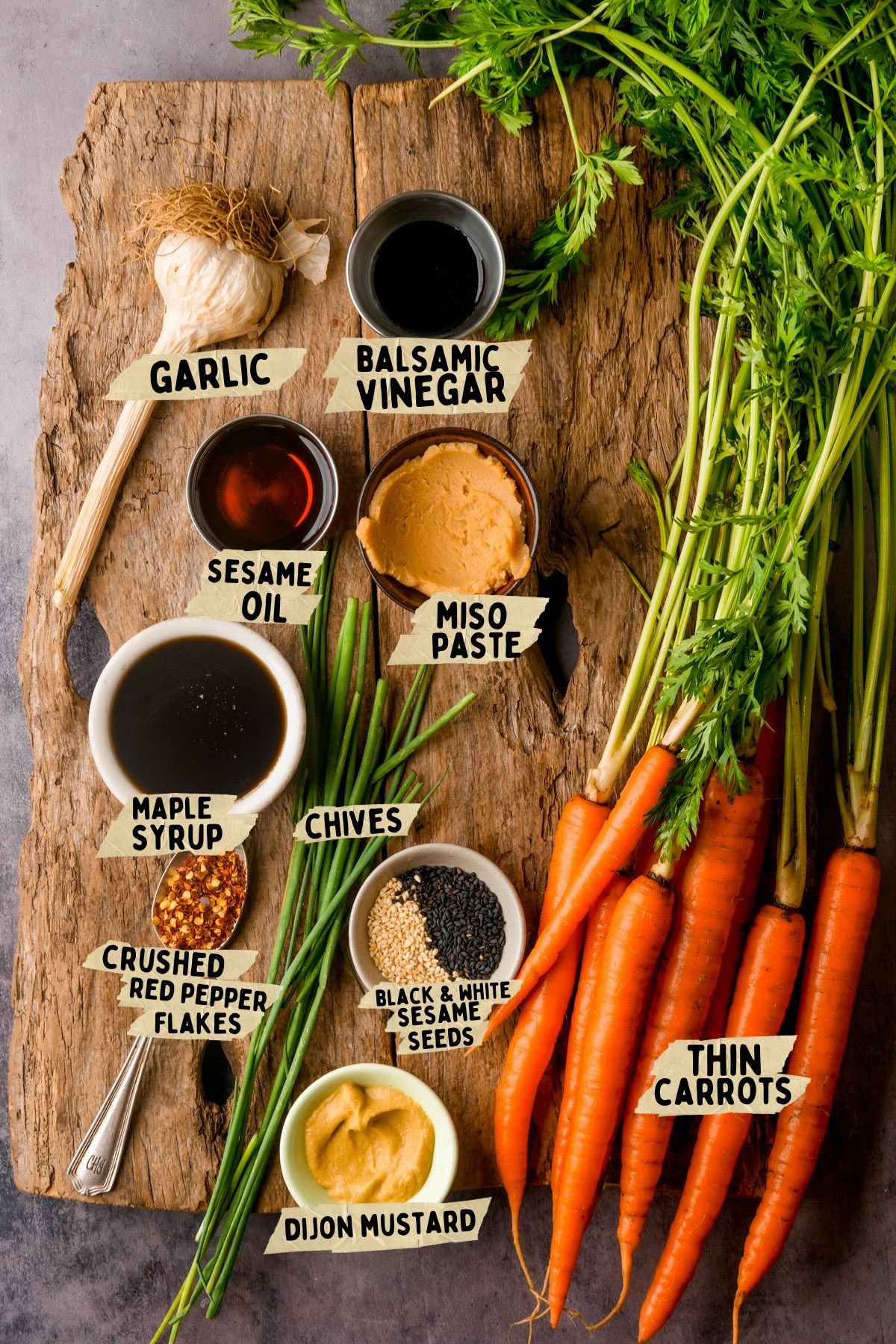A rustic wooden board displays labeled ingredients for a maple roasted carrots recipe: garlic, balsamic vinegar, sesame oil, maple syrup, miso paste, chives, crushed red pepper flakes, black and white sesame seeds, Dijon mustard, and thin carrots with greens.