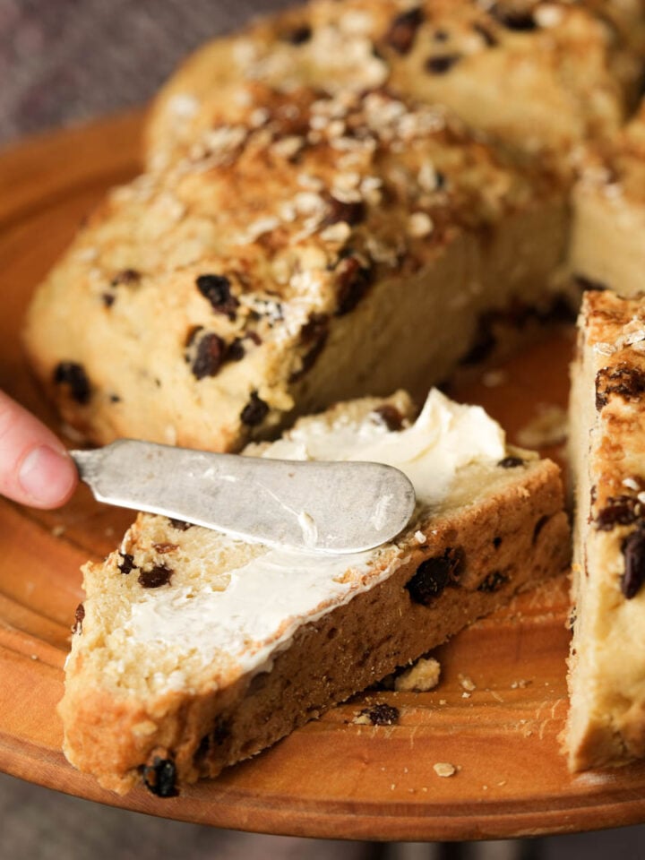 A hand spreads butter on a thick slice of oat and raisin bread using a butter knife. The bread is on a wooden plate with more slices visible in the background.