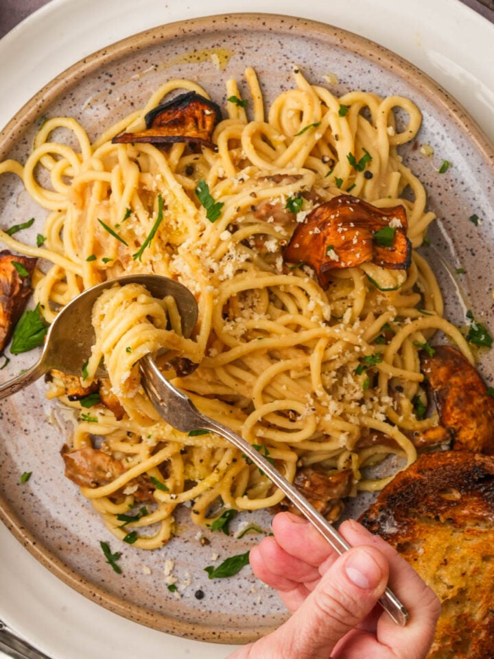 A hand uses a fork and spoon to twirl vegan carbonara with eggplant and parsley on a speckled plate, served with a slice of toasted bread.