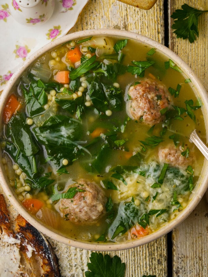 A bowl of vegan Italian wedding soup with meatballs, spinach, pasta, carrots, and parsley sits on a rustic wooden table next to buttered toast, a napkin, and floral salt and pepper shakers.