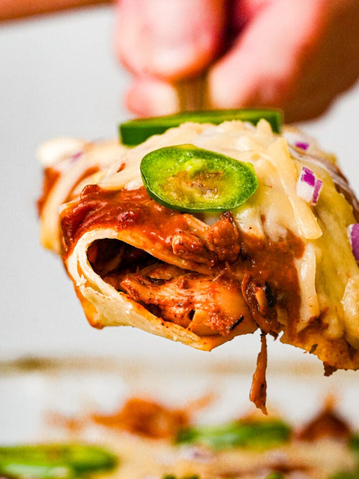 A close-up of a cheesy jackfruit enchilada filled with shredded jackfruit, topped with sliced jalapeños, red onions, and melted cheese, being lifted from a pan by a hand.