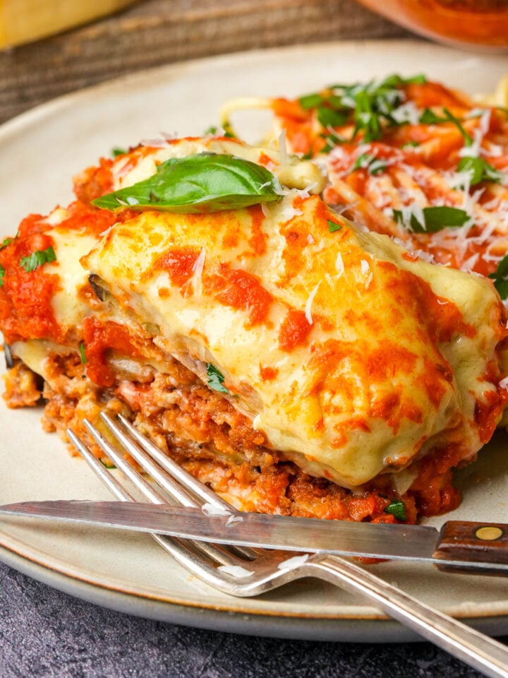 A close-up of a plate with cheesy vegan eggplant parm topped with fresh basil and marinara sauce, next to spaghetti garnished with parsley and grated cheese; a fork and knife rest beside the food.
