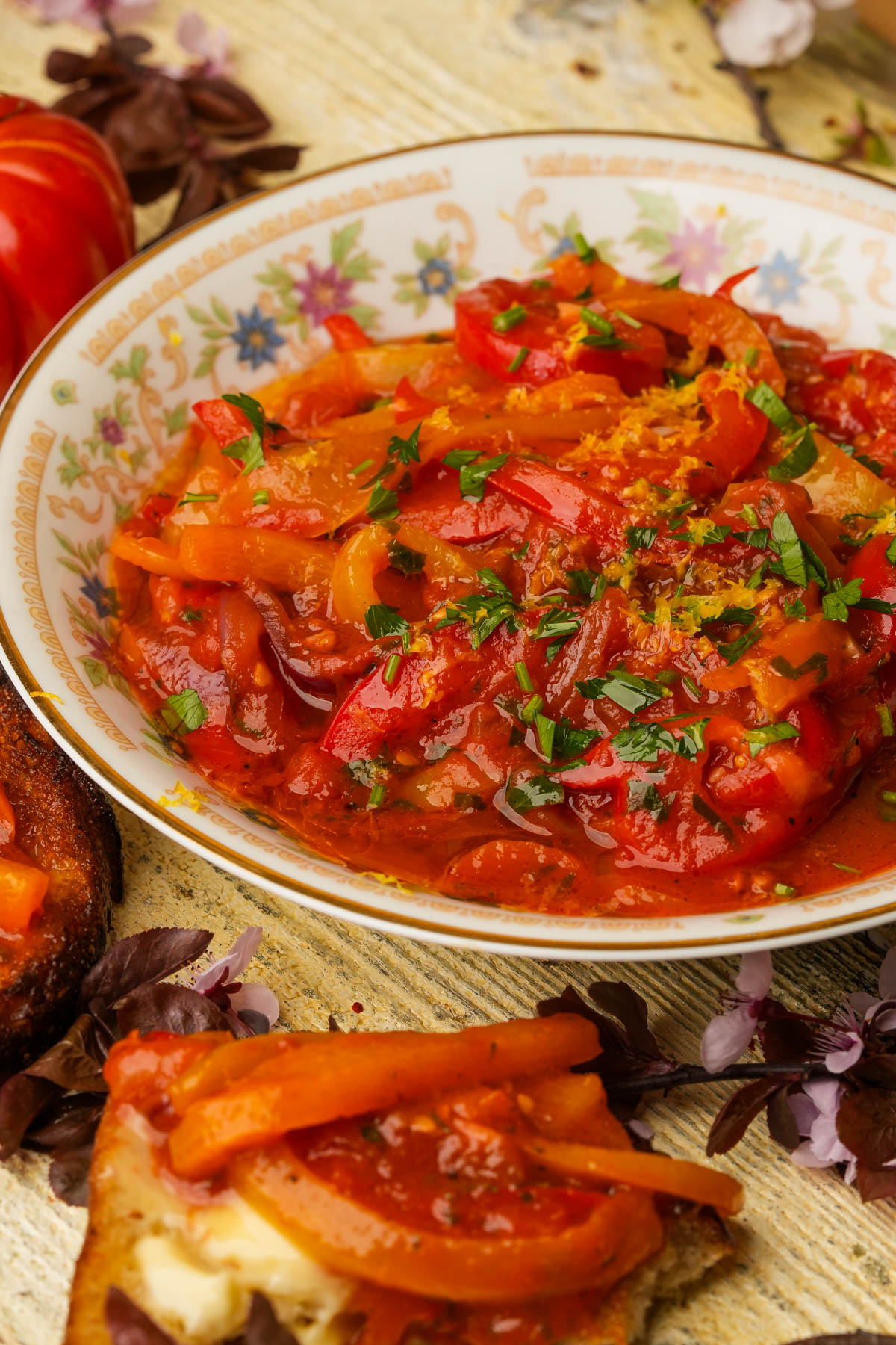 A floral-patterned plate holds peperonata garnished with chopped herbs. Toast with cheese and vegetables sits nearby on a rustic wooden table.