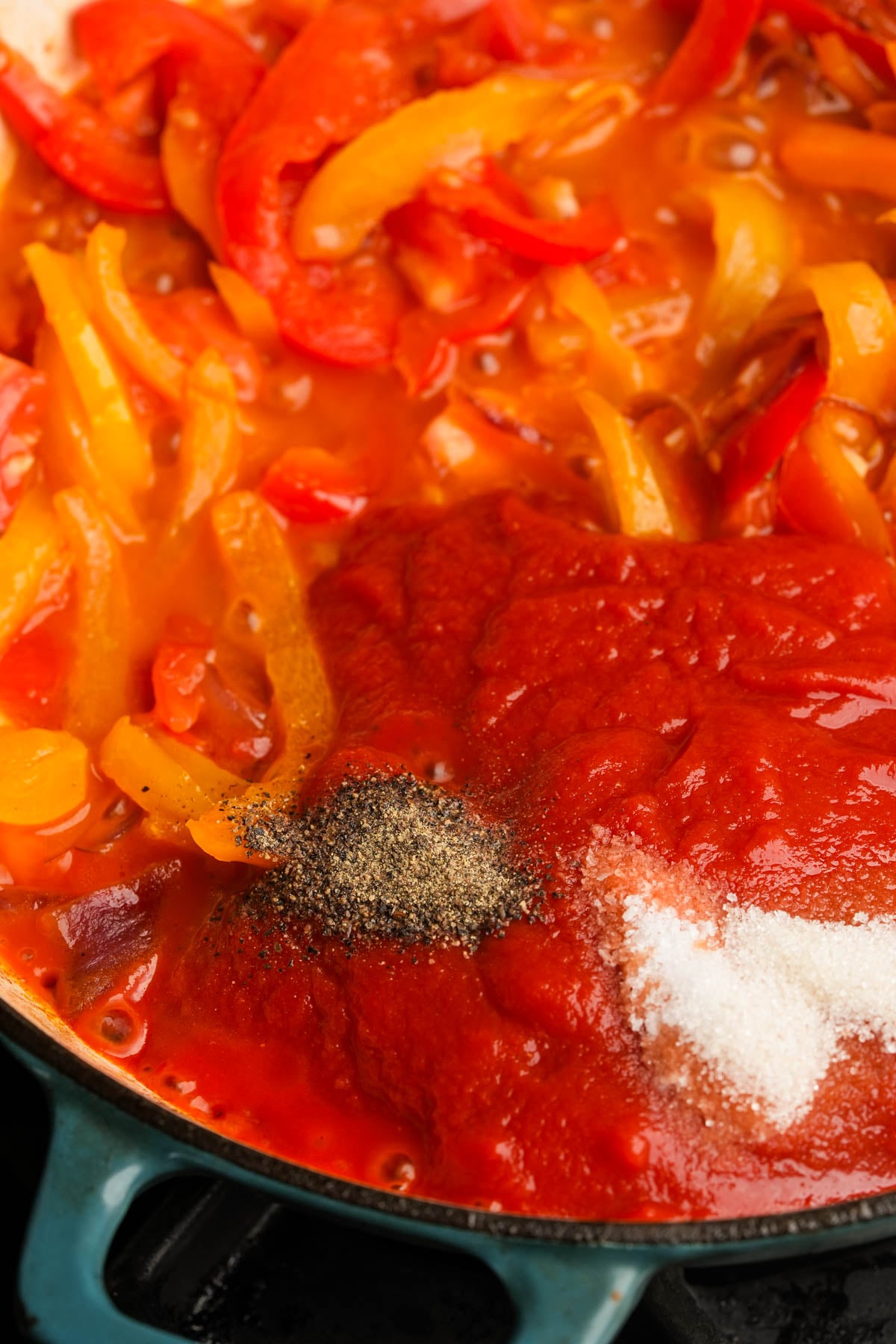 Tomato puree with red and yellow bell pepper strips simmers in a pot, with added salt, pepper and sugar visible on top, ready to be mixed in.